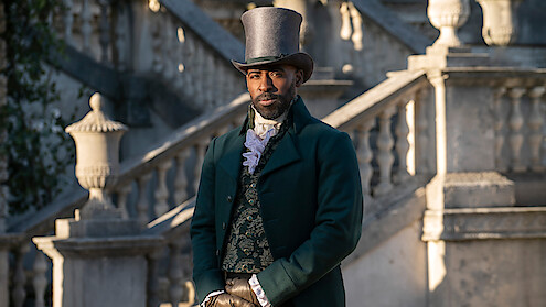 A man in Victorian-era formal attire and top hat stands outside in front of elegant stone steps and balustrades, with classic architecture in the background.