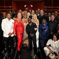 A diverse group of people, including men and women dressed in formal and stylish attire, pose together on a stage in a warmly-lit theater environment, suggesting a celebratory or entertainment industry event.