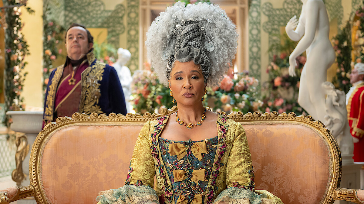 Woman in elaborate 18th-century costume and white wig sits on ornate gold sofa in a lavish, floral-decorated room; man in historical attire stands behind; statue and flowers in background.