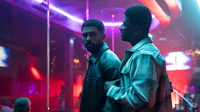 Two men standing together inside a neon-lit nightclub with a pole and colorful lights, both looking serious; background shows other people seated and vibrant decor.