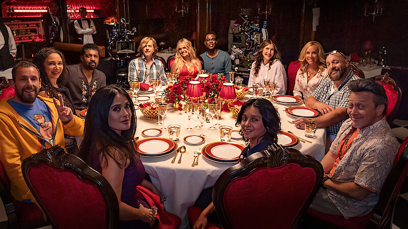A group of people sit around a round dining table set with plates and glasses in an elegant, dimly lit room with red velvet chairs, festive floral centerpiece, and a cozy, celebratory atmosphere.
