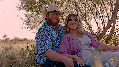 Smiling couple sitting together outdoors in a grassy field, with the man wearing a cowboy hat and patterned shirt, and the woman in a light dress and floral shawl. Sunlit trees and warm, natural light in the background.
