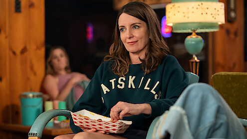 Woman sitting in a cozy wood-paneled room, wearing a sweatshirt and holding a food tray, with another woman blurred in the background and a vintage lamp on a side table.