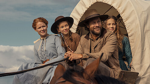A pioneer family sits together in a covered wagon under a blue sky. The man is steering, and three women in period clothing are smiling beside him, capturing a western frontier setting.