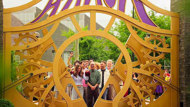 A group of people smiling and posing together behind a large decorative gate with gears and the word "Amuck" above them, set outside with trees and buildings in the background.