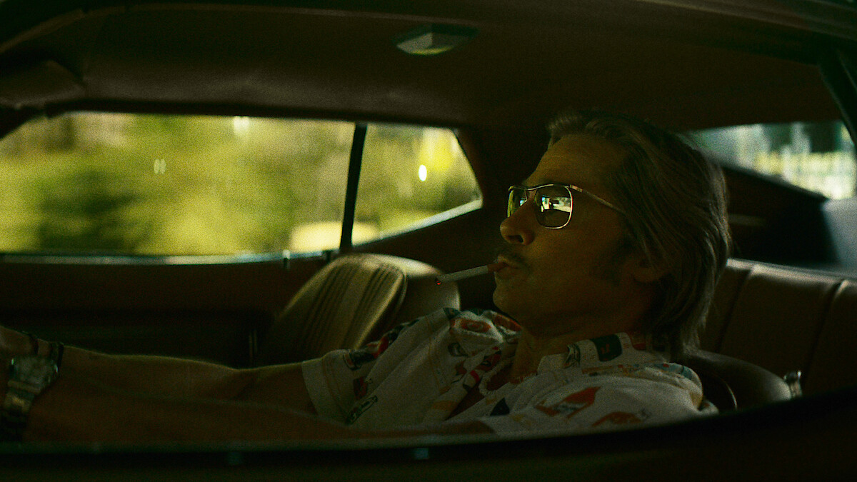 Man wearing sunglasses sits in the driver’s seat of a car, viewed from the side window, with sunlight filtering in. The car interior has a retro style and relaxed vibe.