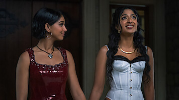 Two young women dressed in formal evening wear stand indoors. One wears a red sequin dress with a heart necklace, the other wears a white corset dress with long hair and shiny makeup. They are holding hands and appear happy.