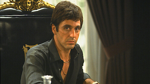 A serious man with short dark hair is seated at a grand, ornate black chair in a dimly lit room with dark curtains and a decorative background, wearing a black shirt and gold necklace.