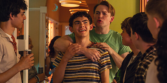 A group of teenagers gather at a lively indoor party. Two boys stand at the center, one smiling while the other playfully wraps an arm around him. The warm, decorated hallway is filled with friends chatting and enjoying themselves.