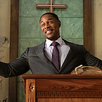 Man in a suit stands at a wooden pulpit with open arms, smiling in a church with a Bible and cross in the background, green and beige walls, and natural lighting.