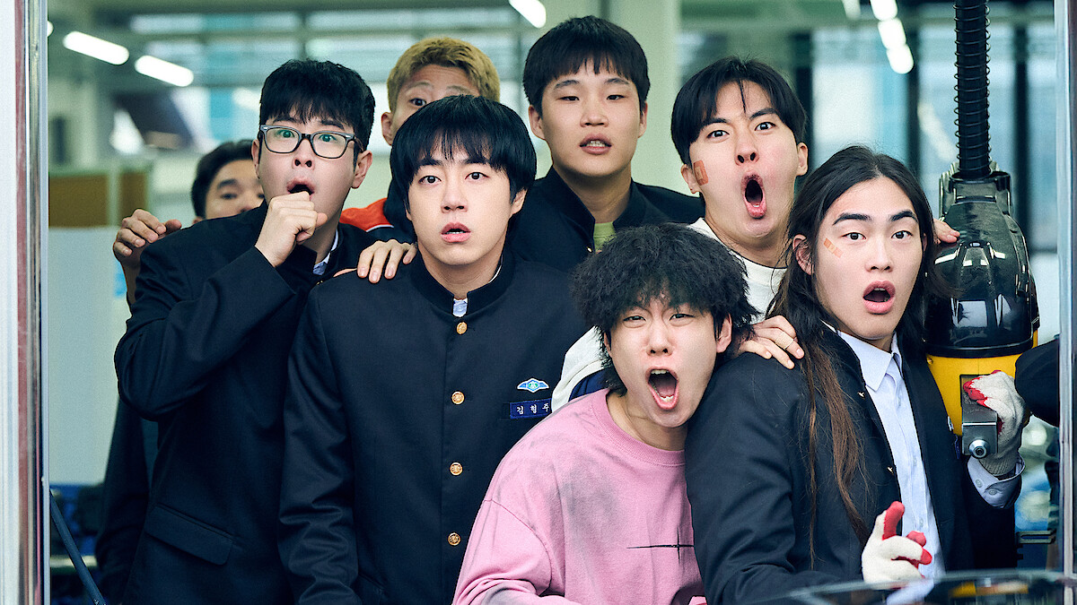 A group of surprised young men in school uniforms and casual clothes stand together indoors, looking in shock at something off-camera in a brightly lit modern setting.