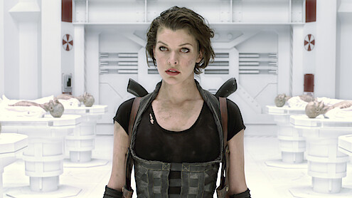 A woman in tactical gear stands in a futuristic, sterile laboratory with white walls and equipment, surrounded by examination tables holding covered bodies, suggesting a sci-fi or action setting.