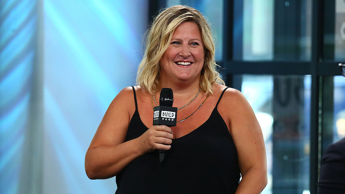 Smiling woman in a black sleeveless top holding a microphone, speaking at a modern indoor event space with large windows, natural light, and a relaxed, positive atmosphere.