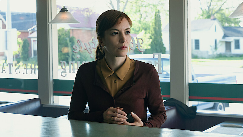 A woman sits alone at a diner counter, looking thoughtful. Sunlight streams through large windows behind her, revealing a suburban neighborhood outside. The diner has a calm, vintage atmosphere.