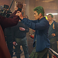 Actor with green hair in a blue shirt poses with a prop sword on a film set, rehearsing a scene with a crew member holding a water bottle; other crew and film equipment visible in the background of an industrial setting.
