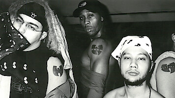 Three men pose in a black and white indoor photo, showing off Wu-Tang Clan tattoos and clothing featuring the group's logo. The environment is casual with urban streetwear and bandanas, emphasizing their affiliation and attitude.