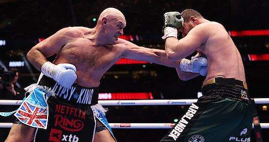 Tyson Fury and Arslanbek Makhmudov in a brightly lit boxing ring, one delivering a punch while the other blocks with his arms up, surrounded by a cheering audience in a large arena.