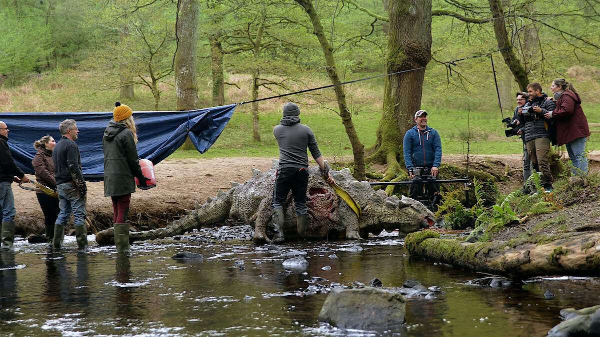 Film crew and technicians work around a dinosaur prop in a forest stream, with cameras and equipment set up for a production scene. The environment is natural, with trees and water, and people wear outdoor clothing.