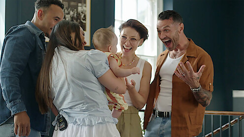 Four adults gathered indoors around a smiling baby, sharing a joyful moment in a brightly lit room with dark walls and framed pictures, one person expressing surprise and excitement.