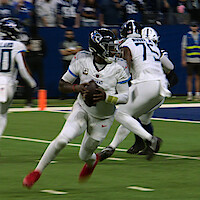 Football quarterback in Tennessee Titans uniform runs with the ball on a field during a game, teammate blocking and opposing players in the background, action shot inside a large stadium.