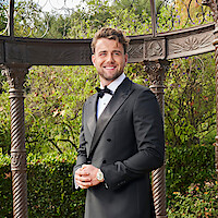 A smiling man in a black tuxedo stands under an ornate metal gazebo in a lush garden setting with greenery and trees in the background.