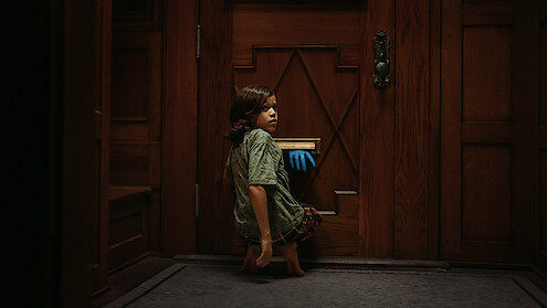 A child crouches in front of a wooden door inside a dimly lit room, looking back as a blue-gloved hand reaches through the mail slot toward them.