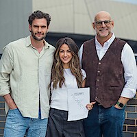 Three people standing outdoors, smiling at the camera. The woman in the middle holds a paper. They are dressed in casual and smart-casual attire, with a modern building structure visible in the background.