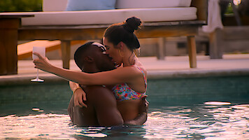 A couple in swimwear passionately kisses while embracing in a pool; the woman holds a drink and the setting appears to be an outdoor luxury resort with lounge chairs in the background.