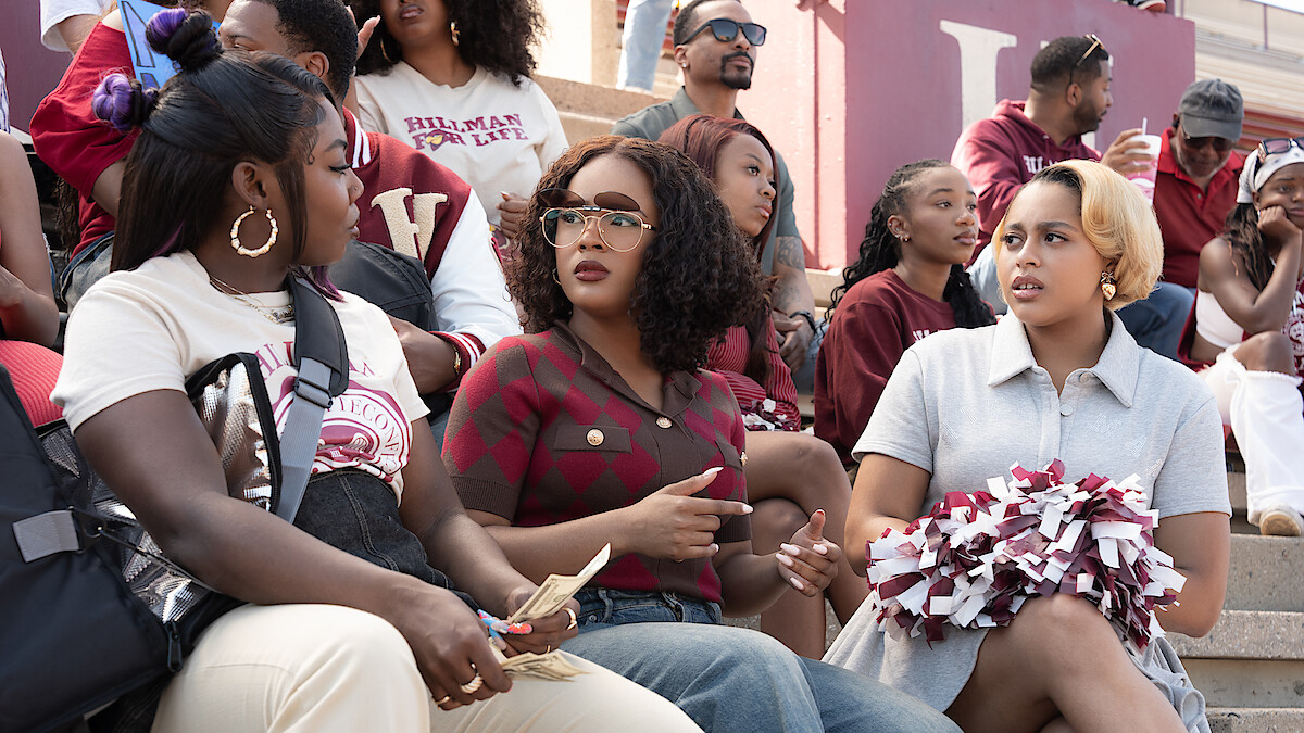 Three women sit on stadium bleachers at a sports event, talking. One holds cash, another has pom-poms. People in the background wear maroon and white, suggesting a school or college environment.