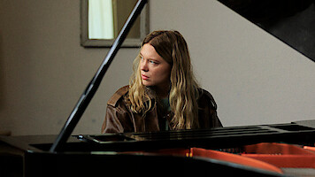 A person with long hair and a brown jacket sits at a grand piano indoors, looking to the side. The setting is softly lit with a neutral wall and a mirror in the background.
