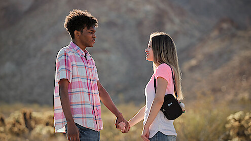 Two people holding hands and facing each other outdoors in a desert landscape with mountains and cactus in the background, wearing casual clothes and appearing to enjoy the natural setting.