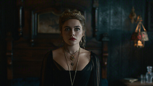 A woman in a dark, ornate dress with layered necklaces stands in a dimly lit, elegant room with vintage wooden furniture and decorative lighting, creating a moody and dramatic atmosphere.