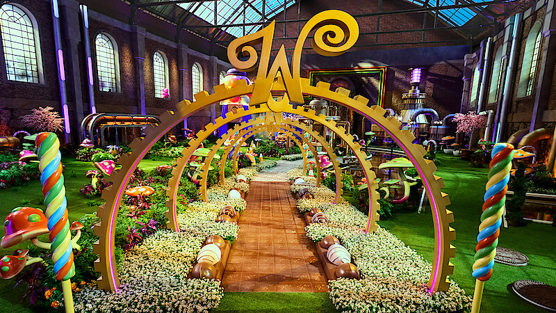 A whimsical candy-themed garden inside a large factory with colorful sweets, oversized mushrooms, lollipops, golden arches, flowers, and a large "W" centerpiece under a glass roof, creating a magical, playful atmosphere.