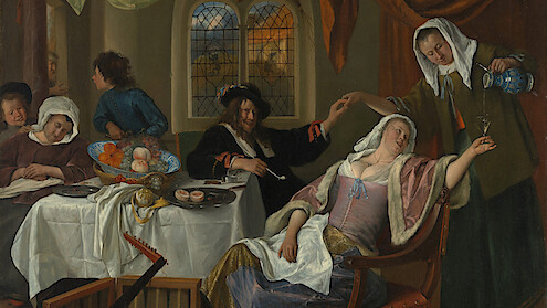 Group of people in 17th-century clothing at a dining table in a richly decorated room; a woman reclines in a chair as another pours her a drink, with food and musical instruments nearby.