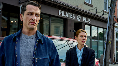 Two serious-looking people stand outside a Pilates studio on a city street, with a car parked behind them; the mood appears tense and contemplative.