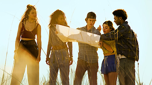 Five young people stand in tall grass outdoors, closely examining a large map together in bright daylight, appearing focused and collaborative in a natural setting.