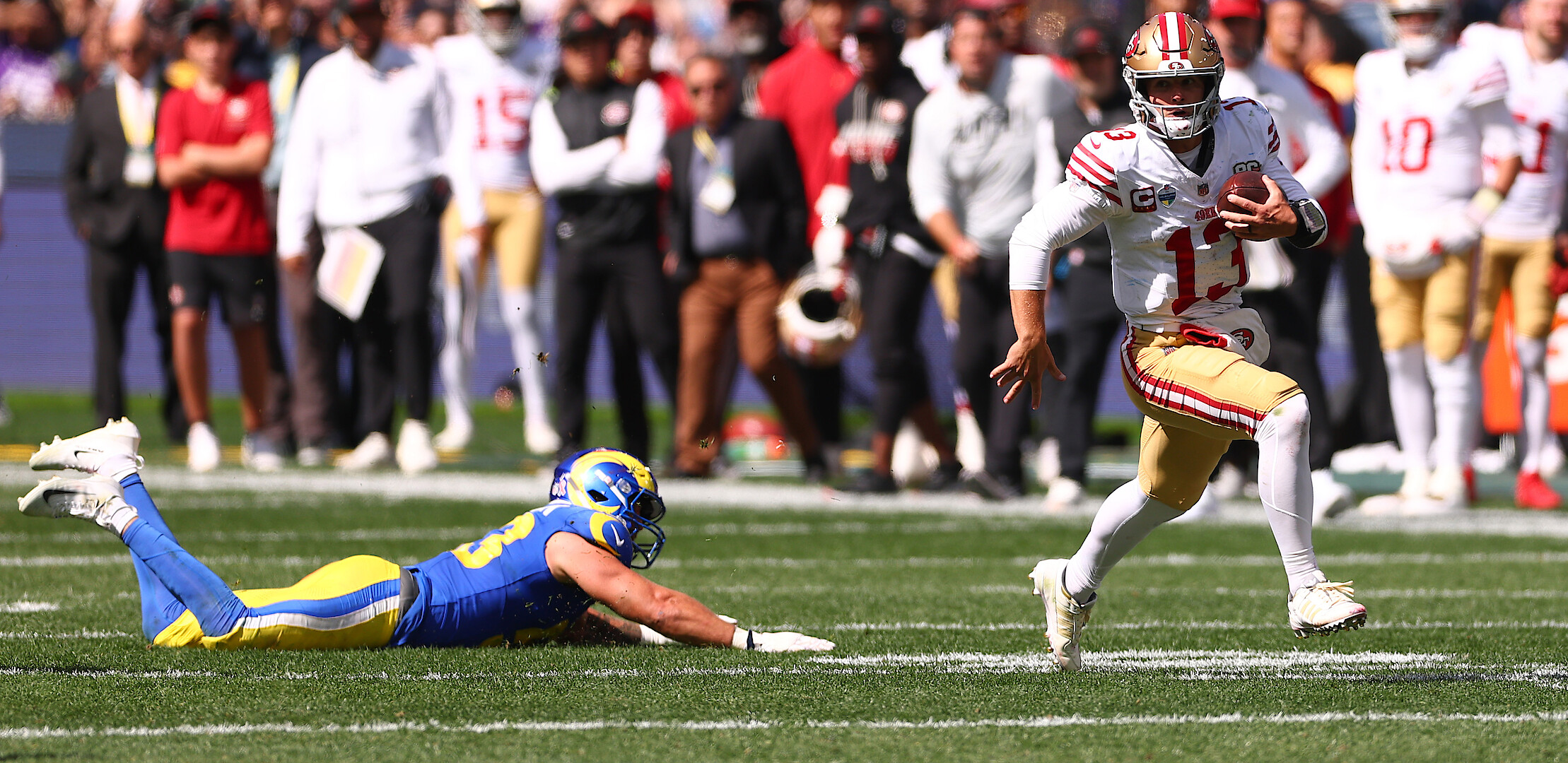 Football quarterback in a white and gold uniform runs with the ball on a sunny field while a defender in a blue and yellow uniform dives to tackle him; crowd and teammates watch in the background.