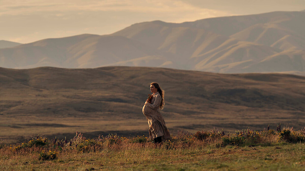 Pregnant woman in a long dress stands in a grassy field with wildflowers, set against a backdrop of rolling hills and mountains during golden hour, creating a serene and natural outdoor scene.