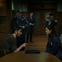 A man and a woman sit across from each other at a table in a dimly lit room, with police officers and investigators working in the background, suggesting an interrogation or police investigation scene.