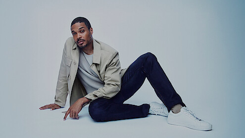 Ray Fisher wears dark jeans and a tan jacket, and sits in a jaunty pose.