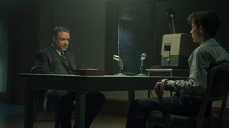 Two men sit across from each other in a dimly lit interrogation room, one appearing to question the other who is attached to a polygraph machine, with microphones and recording equipment on the table.