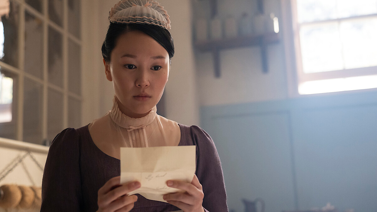 Woman in period costume reading a letter in a softly lit, vintage room with shelves, jars, and a window in the background.