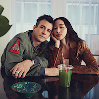 Um homem e uma mulher estão sentados próximos um ao outro, apoiados em uma mesa de vidro em um ambiente iluminado com grandes janelas. Há um suco verde sobre a mesa e um vaso de flores ao canto, criando uma atmosfera aconchegante e intimista.