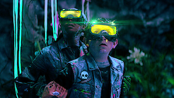 Two people wearing yellow glowing goggles and denim jackets with patches stand in a colorful, neon-lit cave environment, surrounded by plants and glowing decorations, looking upward as if intrigued or cautious.