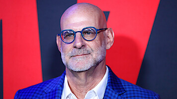 Bald man with glasses and a beard wearing a blue checkered jacket and white shirt, standing in front of a red and black event backdrop.