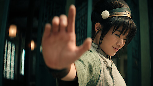 Miyako as Toph, with long hair in a bun, wearing a headband and traditional attire, smiling gently, extends hand outward confidently.