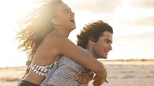 Two young adults enjoying a sunny day at the beach, with the woman laughing as she rides piggyback on the man, both appearing happy and carefree, with sand and bright sky in the background.