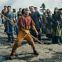 A group of people in costumes stand outdoors on rocky terrain with trees and mountains in the background, the central figure holding a staff and looking alert.