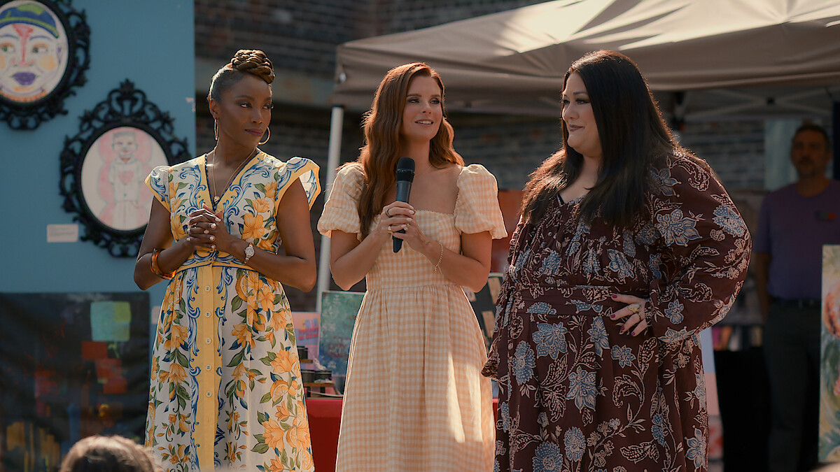 Three women in summer dresses stand together outdoors at an art fair or festival, one holding a microphone. Colorful paintings and a canopy tent are visible in the background.