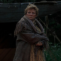 A woman in worn clothing and a scarf stands outdoors in front of a rustic tent, surrounded by greenery, with a serious expression and arms crossed in a natural, forested setting.